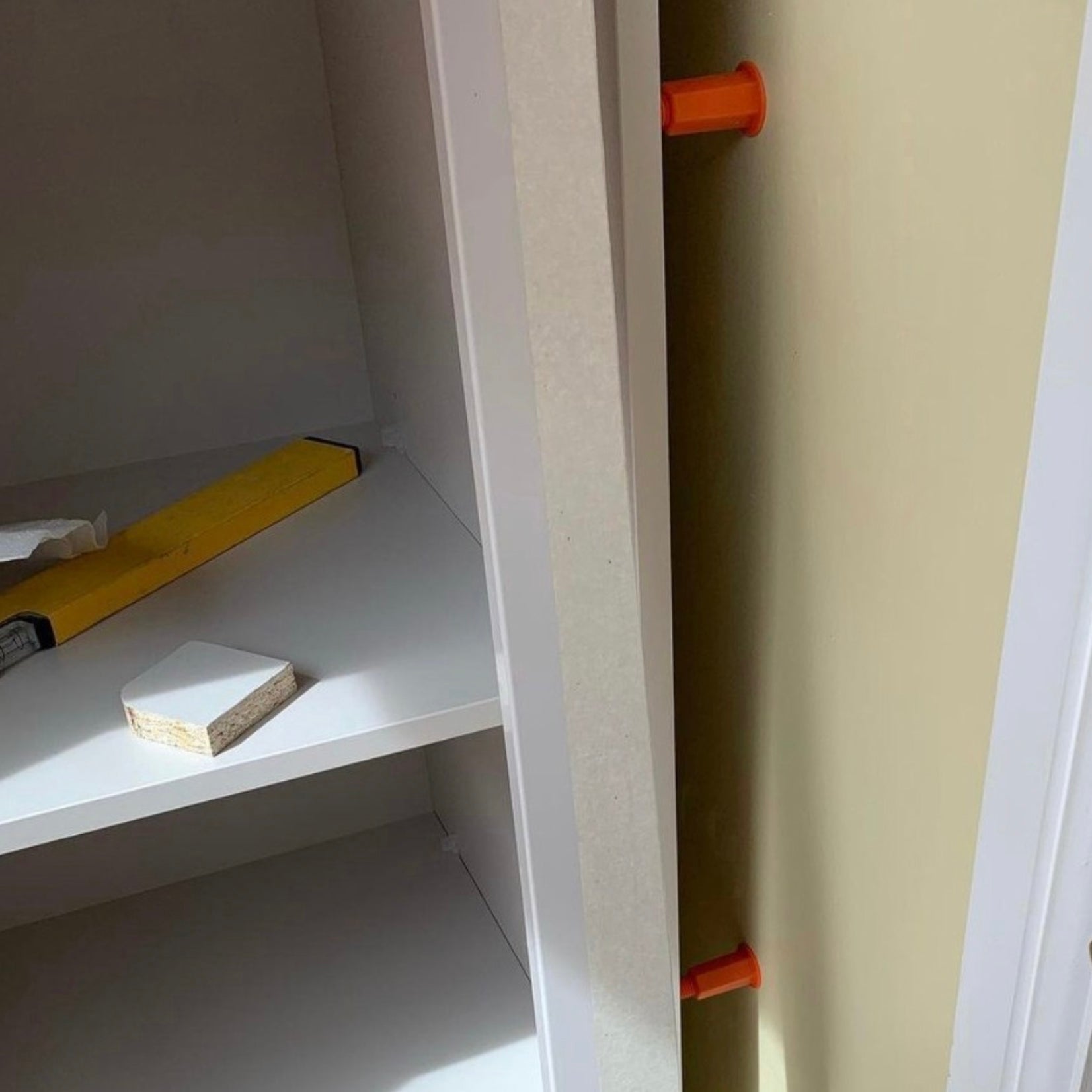 Étagère blanche avec fixations réglables Space-Plug orange insérées verticalement le long de la face intérieure du meuble, présentée dans un environnement intérieur avec un niveau à bulle jaune et de petites cales posées sur l'étagère.